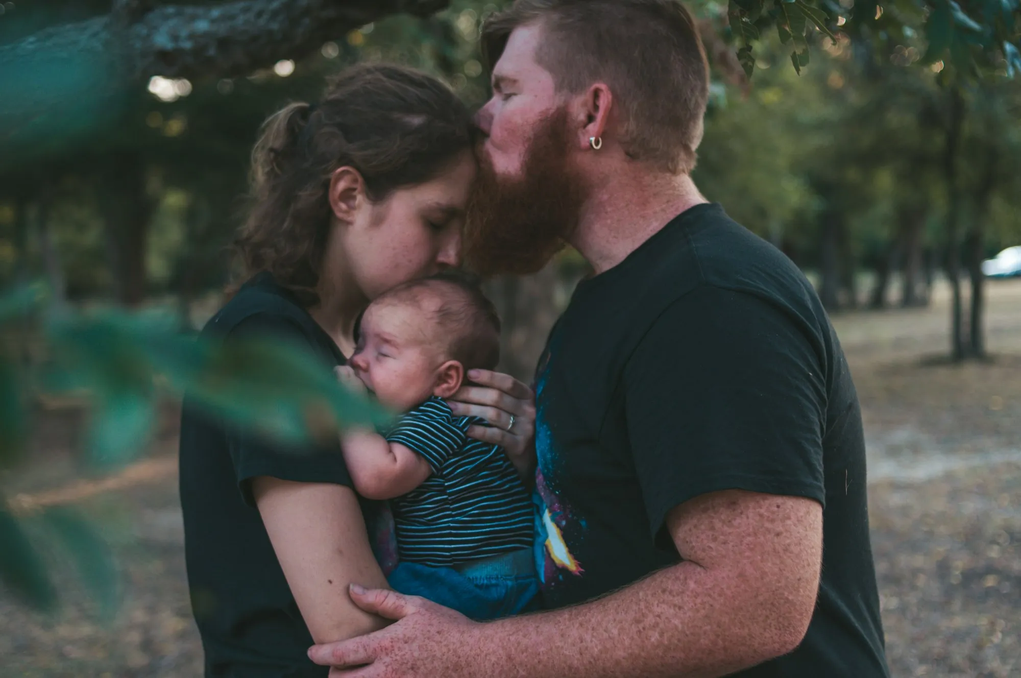 Bearded man kissing his partner's forehead while she holds their baby outdoors