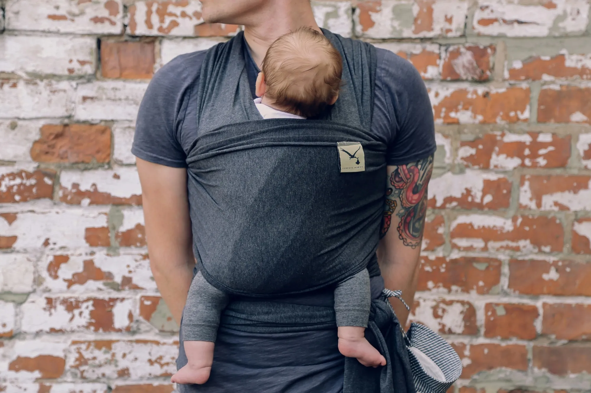 Dad carrying his baby in a dark gray wrap carrier, standing in front of a whitewashed brick wall