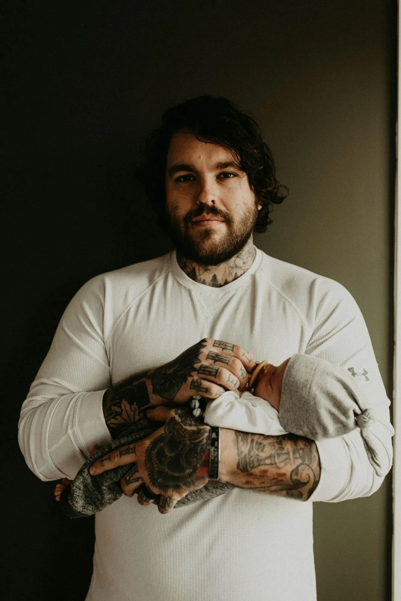 Bearded, tattooed dad in a white sweater cradling his sleeping newborn in a knotted gray hat
