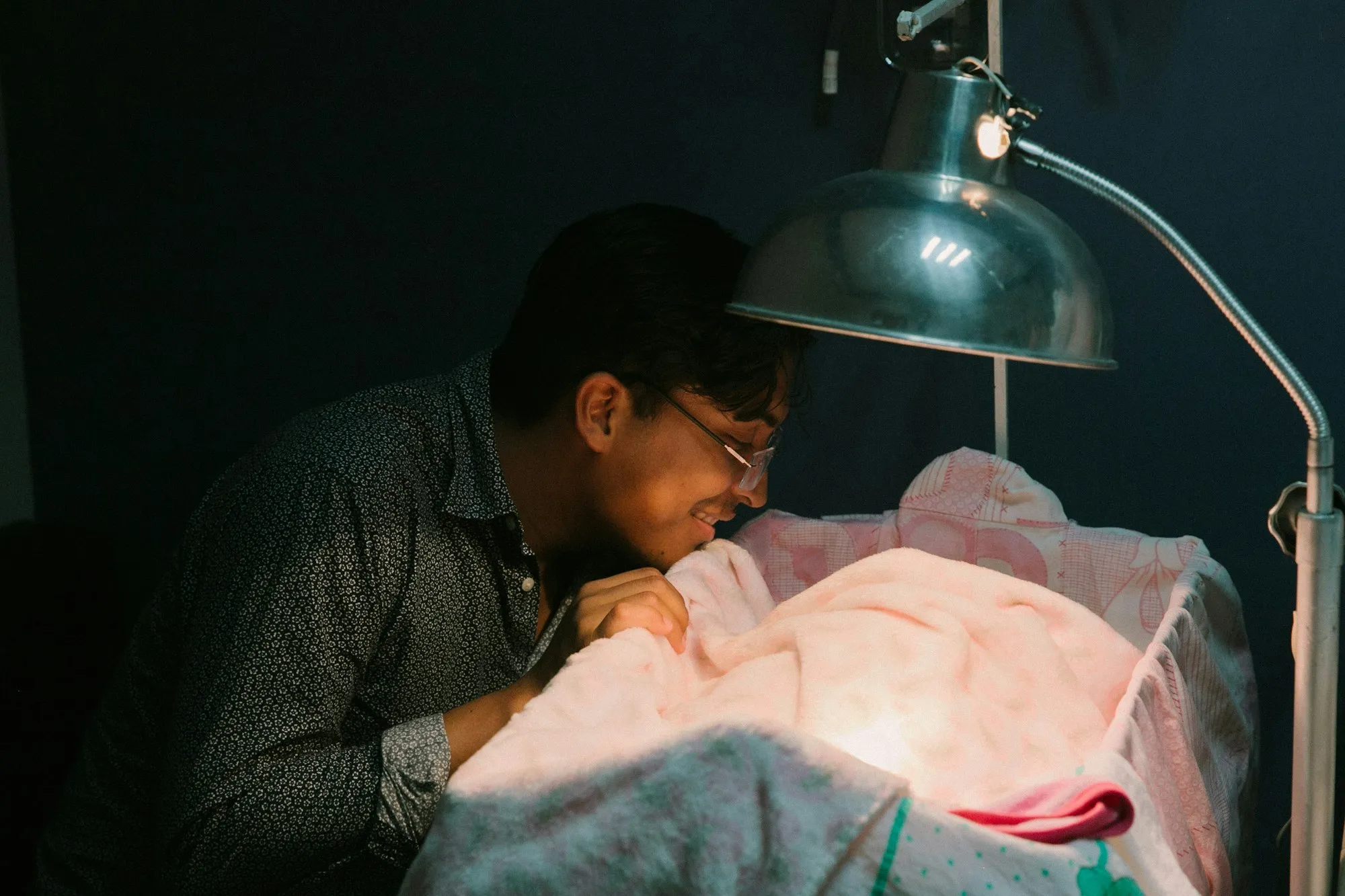 Dad leaning over a hospital bassinet under a lamp to look at his newborn