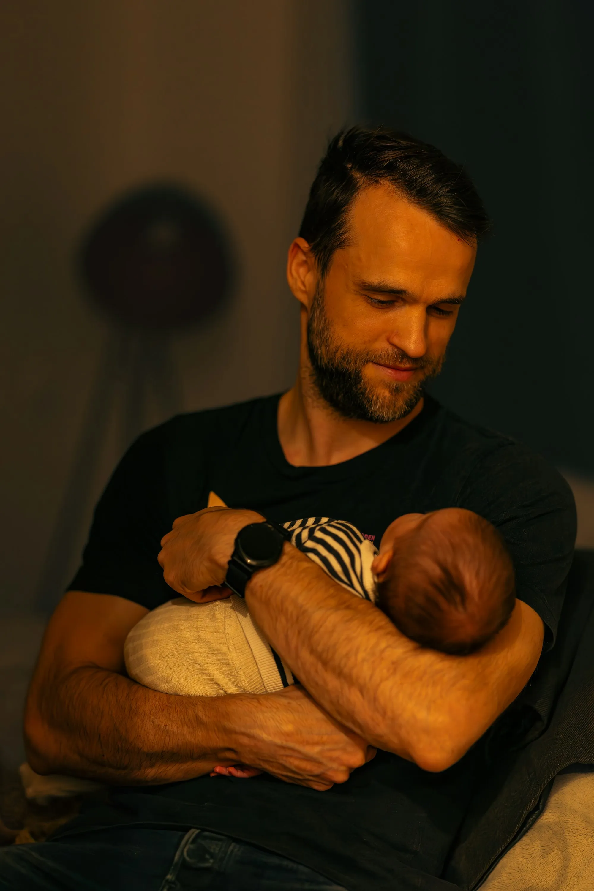 Dad in a dim room in the evening, smiling down at the newborn in his arms