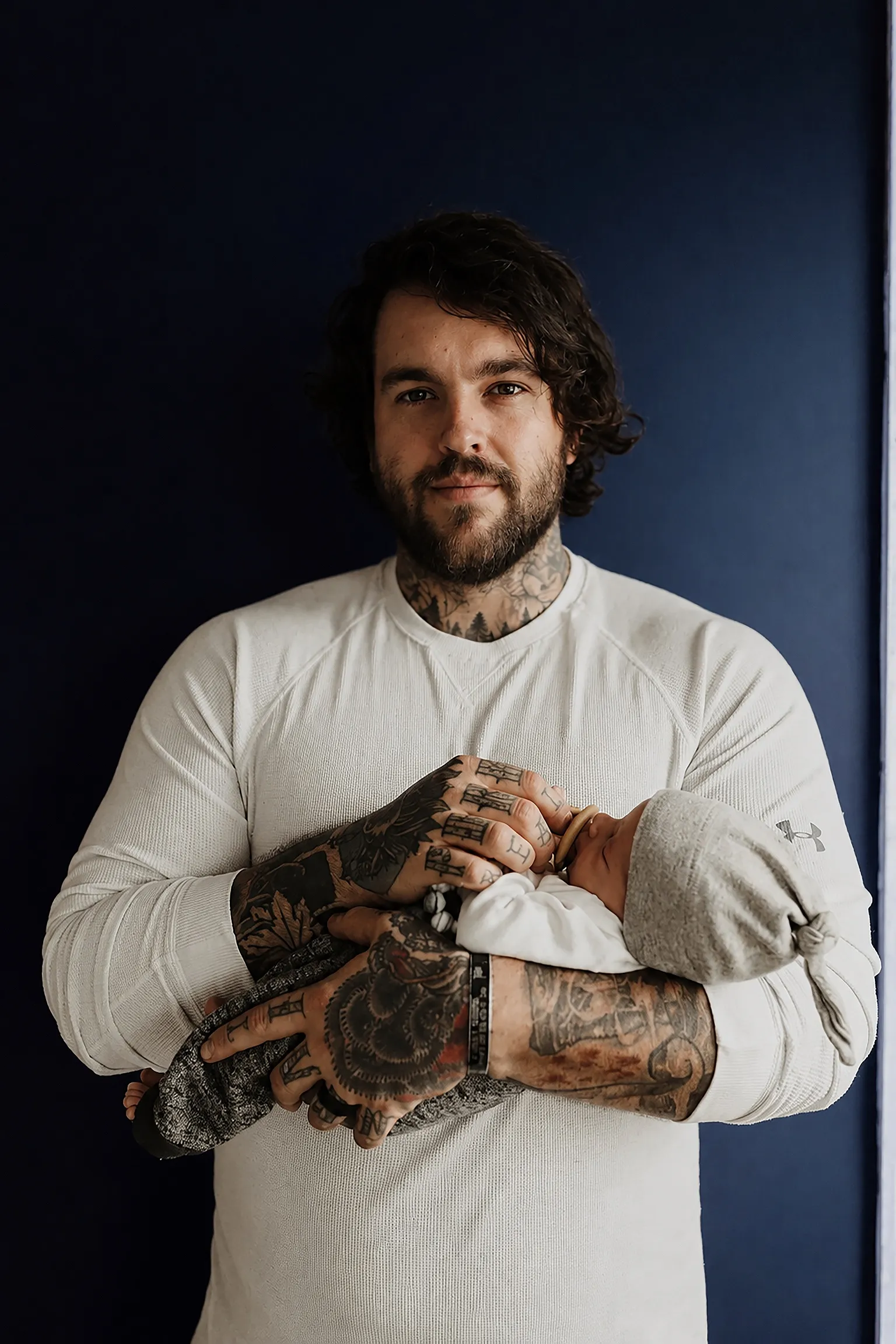 Bearded, tattooed dad in a white sweater holding his sleeping newborn against a dark blue wall