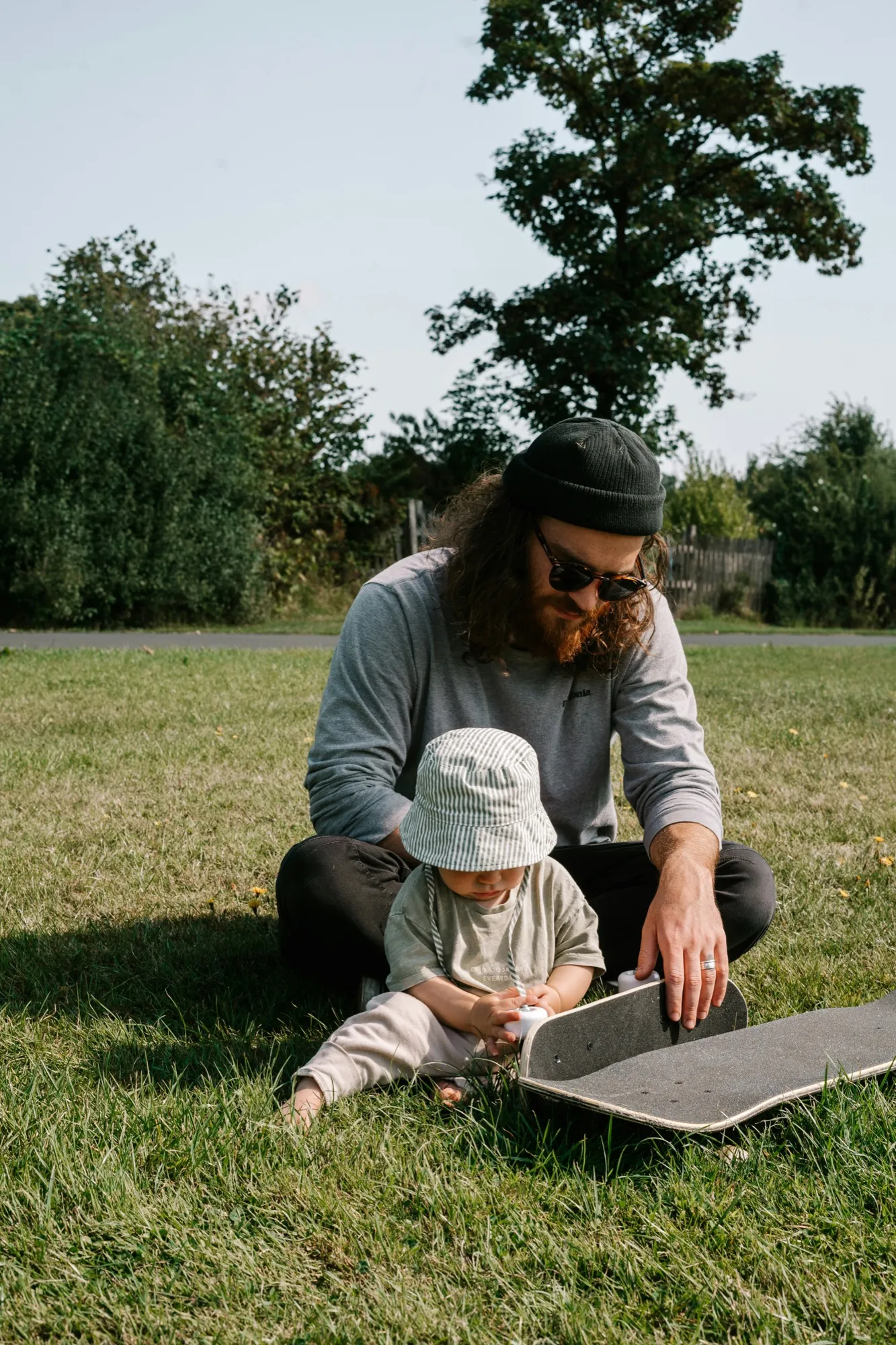 Dad in a beanie sitting on the grass with his toddler in a sun hat