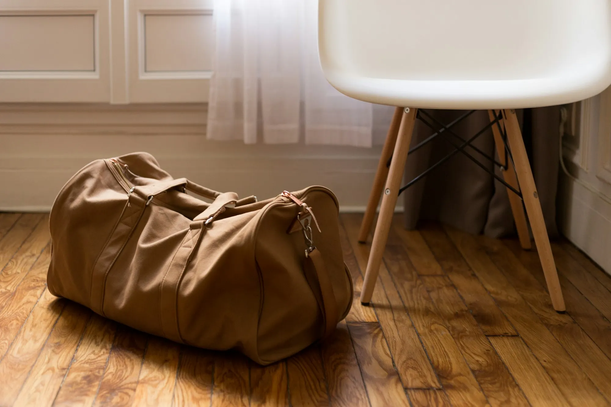 Packed brown duffel bag on a wooden floor next to a white chair