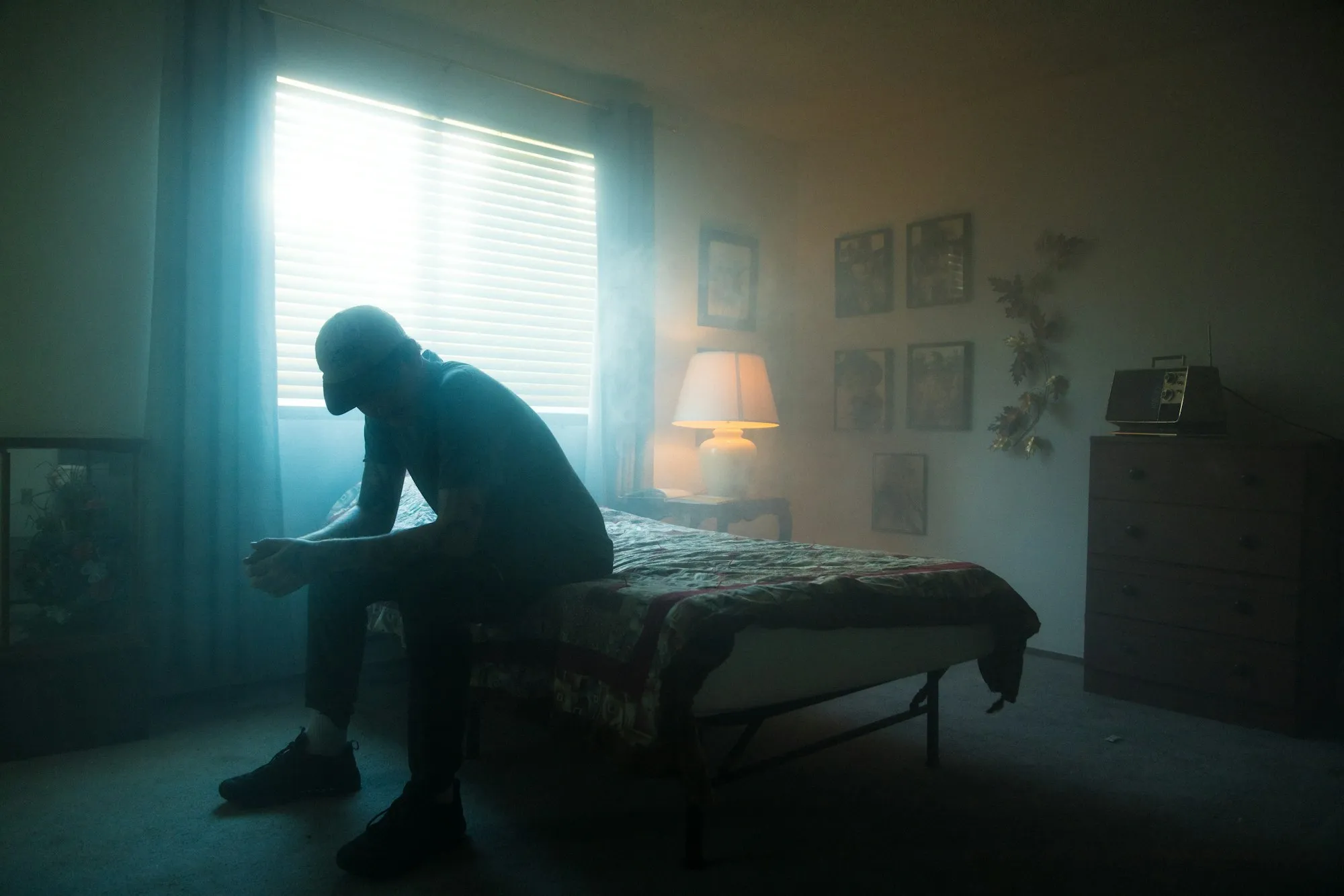 Man sitting on the edge of a bed with his head down, light coming through the blinds