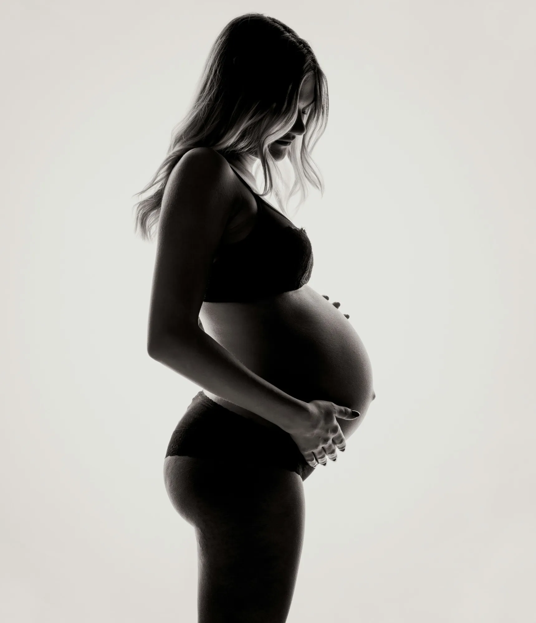 Pregnant woman in silhouette, looking down and holding her belly with both hands