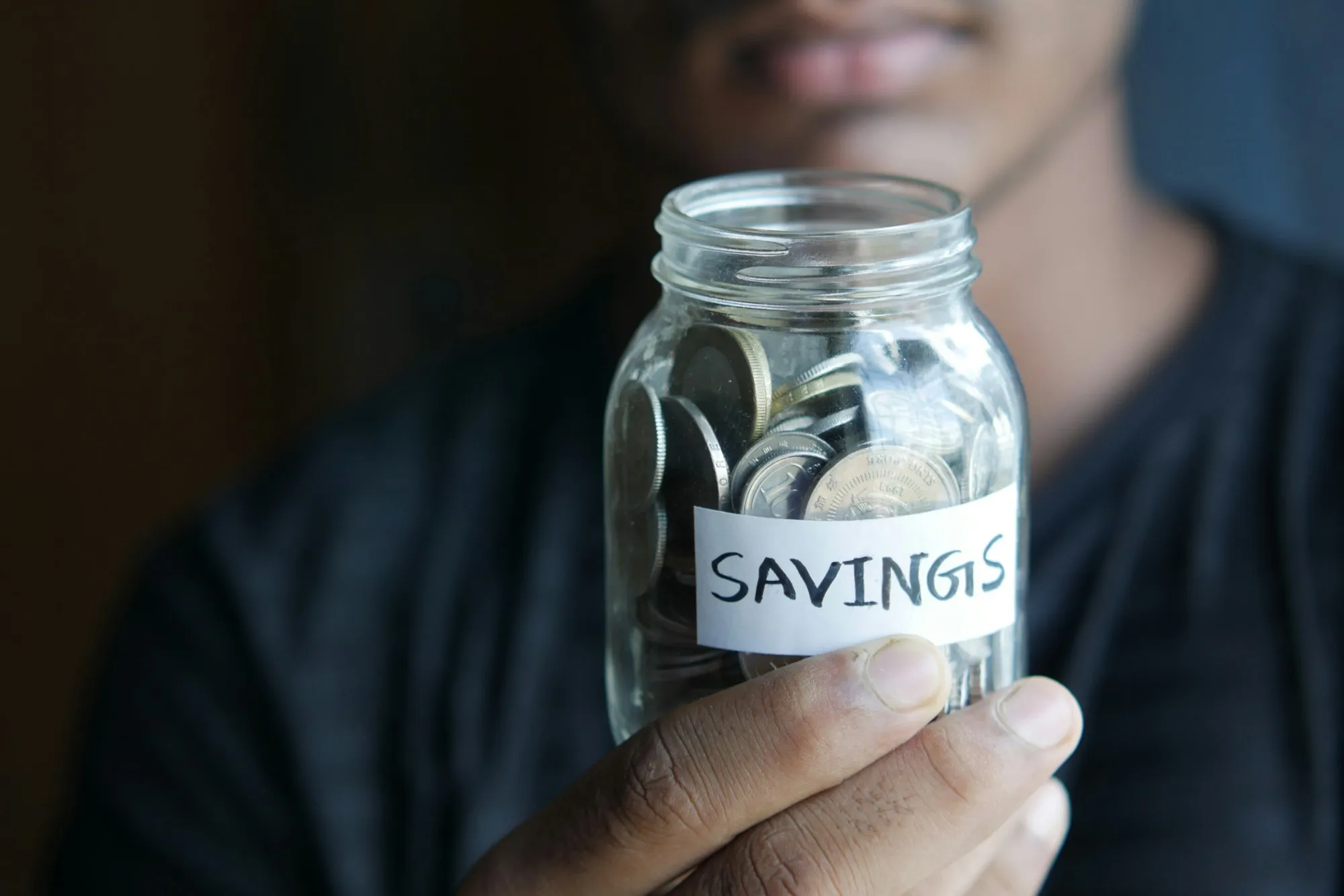 Hand holding a glass jar of coins labeled Savings
