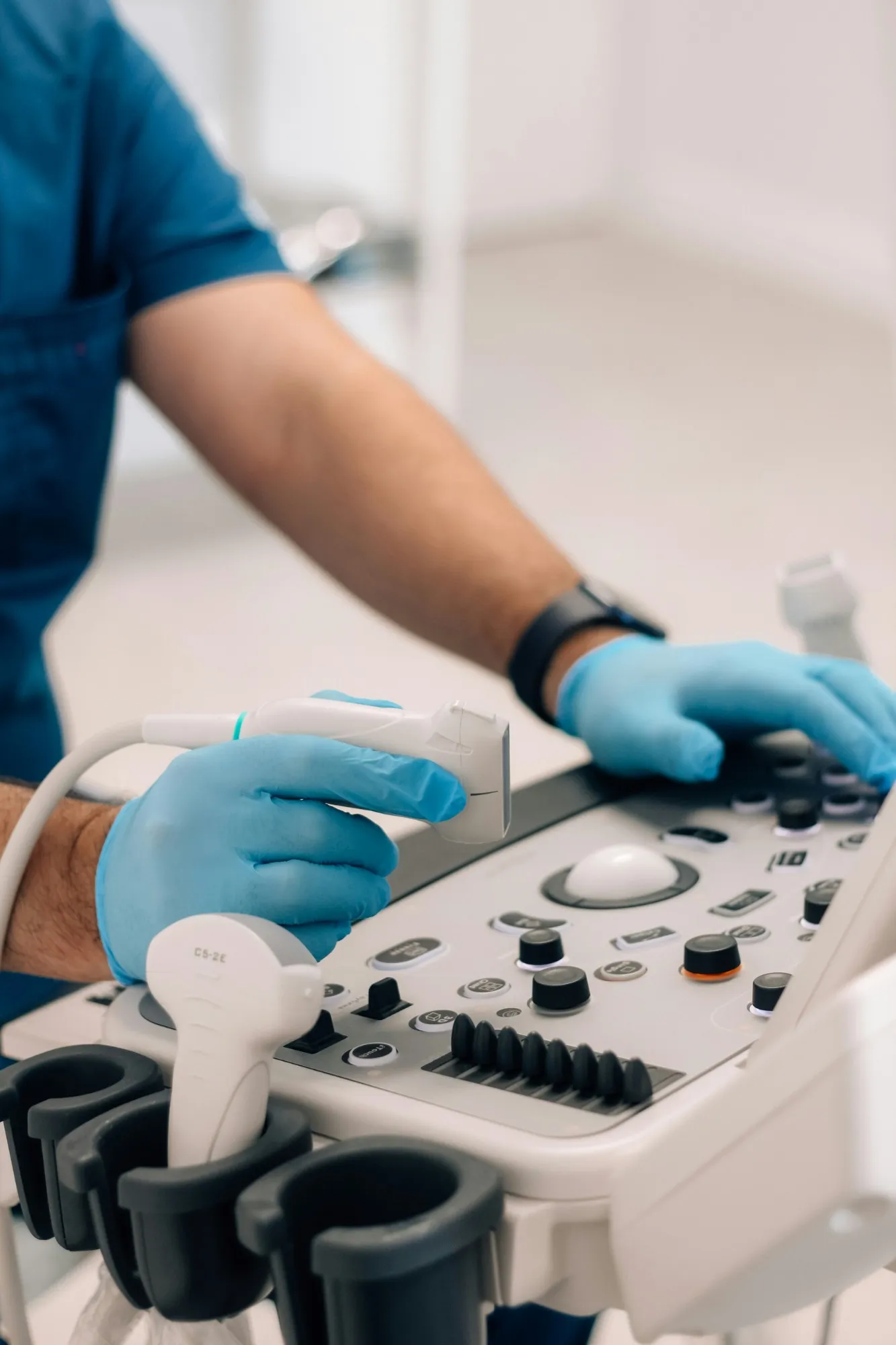Sonographer in blue gloves holding an ultrasound probe over the controls of an ultrasound machine